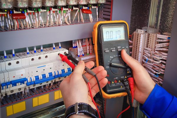 Multimeter is in hands of electrician on background of electrical automation cabinet. Adjustment of electrical control circuit for industrial equipment.